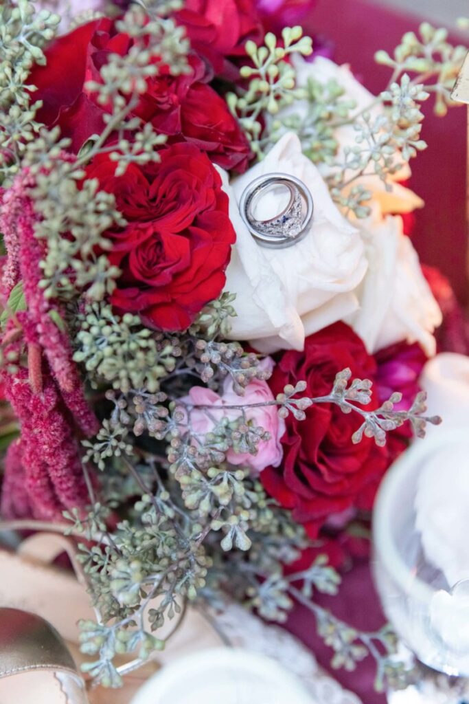 Shannon-James-Wedding-Low-Res-96 - TOMMY GATZ ENTERTAINMENT Bouquet of red and white roses with silver rings nestled among them, surrounded by greenery and pink flowers.