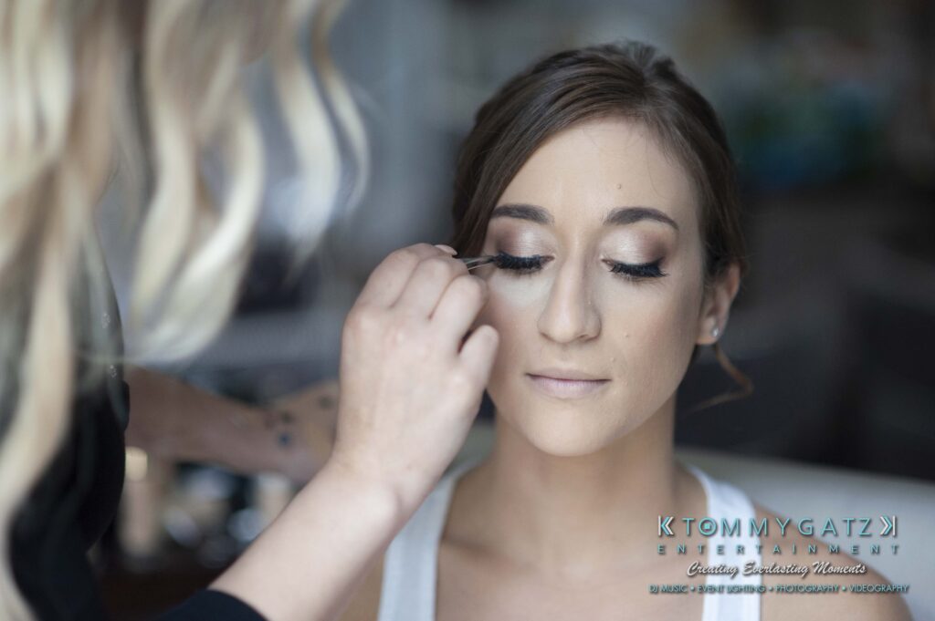 A woman is having her makeup done with a focus on her eyes. She has her eyes closed and is wearing a white top. Another person is applying the makeup.
