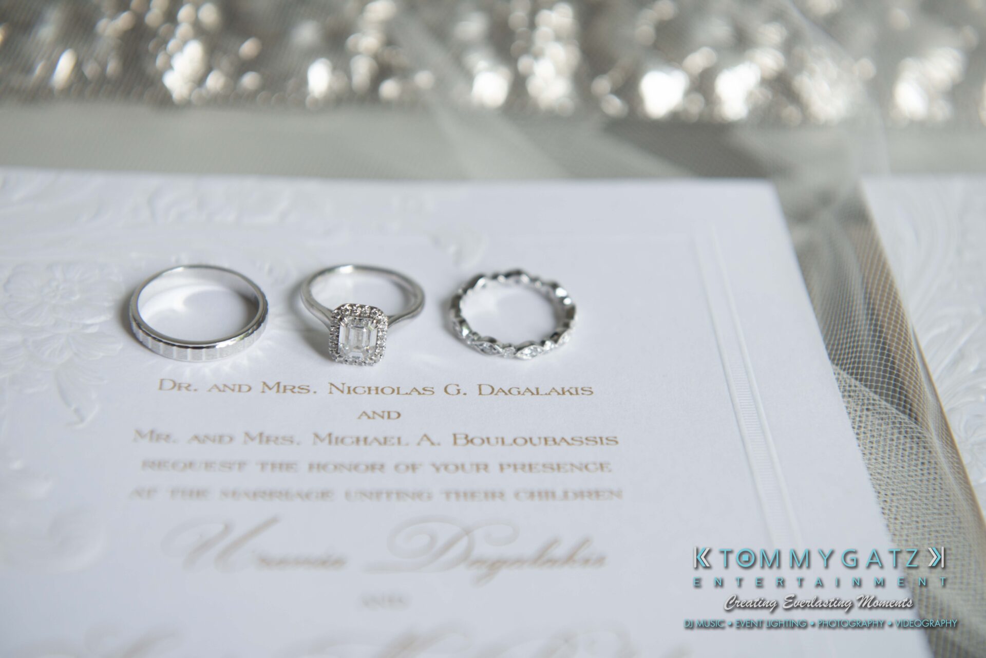 Urania-Aristidis-060218-PREP-Low-Res-WM-18 - TOMMY GATZ ENTERTAINMENT Three rings, including a diamond ring, rest on a wedding invitation with embossed floral details. A logo for Tommy Gatz Entertainment is visible at the bottom right.