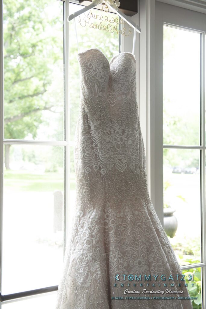 A strapless, lace-covered wedding dress hangs by a window, with a personalized hanger that reads "June 2, 2018.