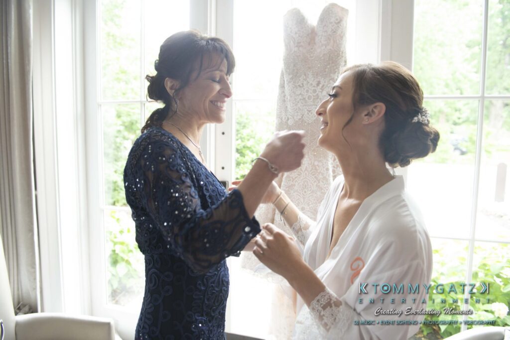 Two women smiling and standing near a wedding dress by a window. One is wearing a navy dress, and the other, in a white robe, has her hair styled up.