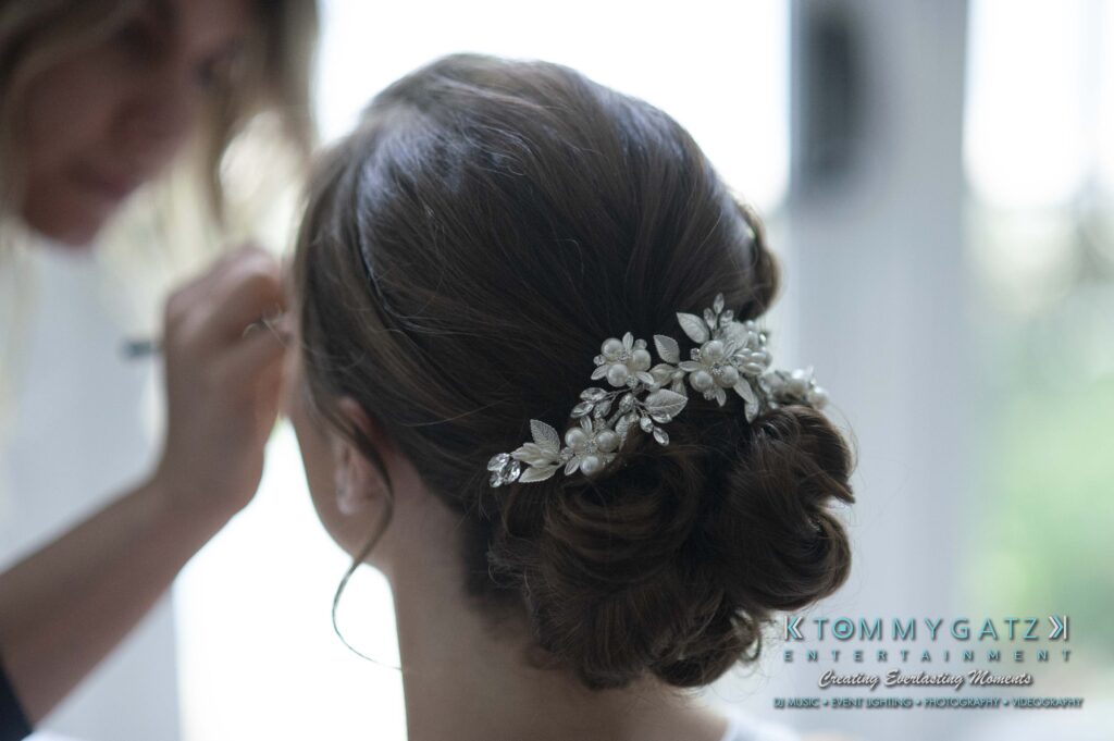 A woman with a floral hair accessory is having makeup applied by another person. Logo reads "Tommy Gatz Entertainment.