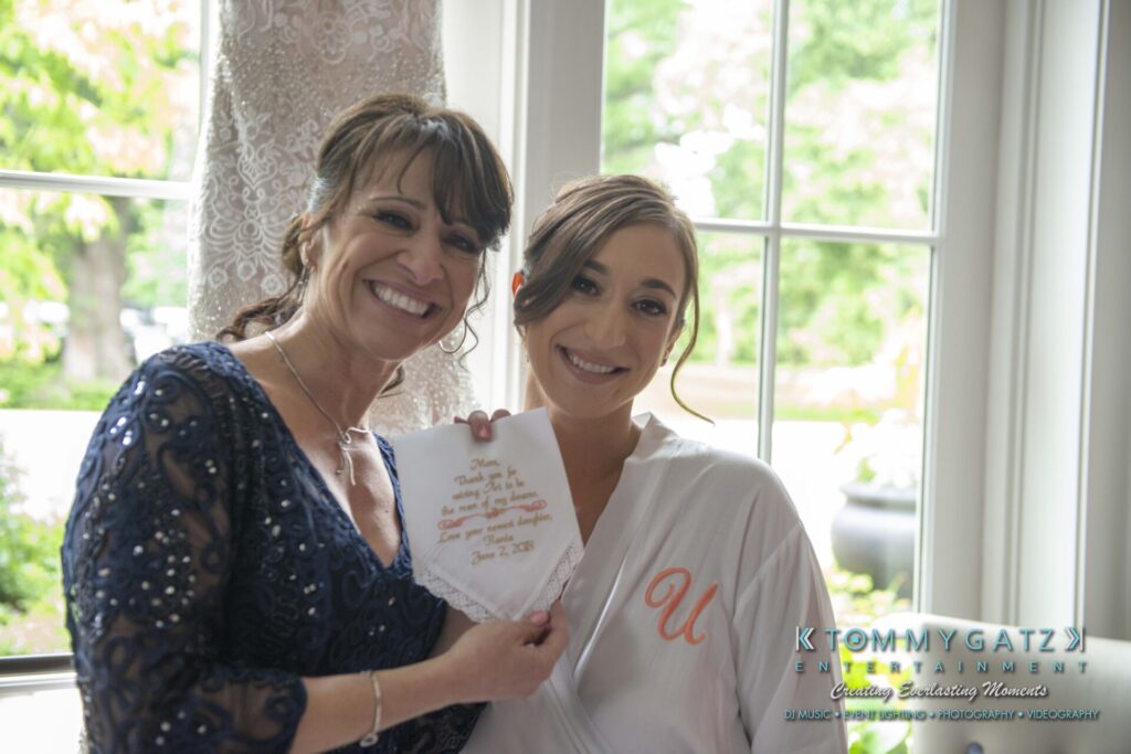 Two women smiling indoors, one holding a handkerchief with stitched text. They stand near a window.