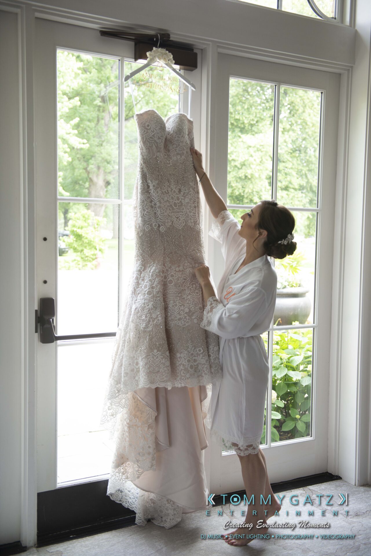 Urania-Aristidis-060218-PREP-Low-Res-WM-48 - TOMMY GATZ ENTERTAINMENT A woman in a robe admires a hanging lace wedding dress by a tall window in a bright room.
