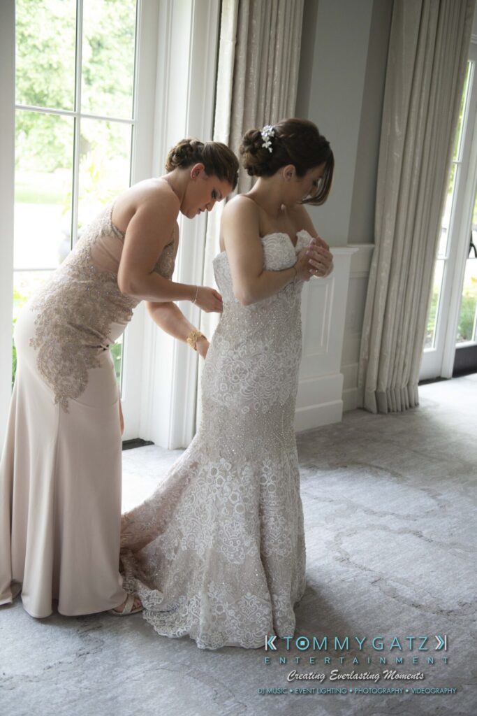 A woman assists another woman in fitting a lacy wedding gown near a window.