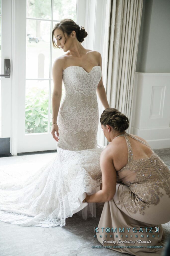A bride stands in a lace wedding gown as another woman adjusts the train. They are indoors, near a large window.