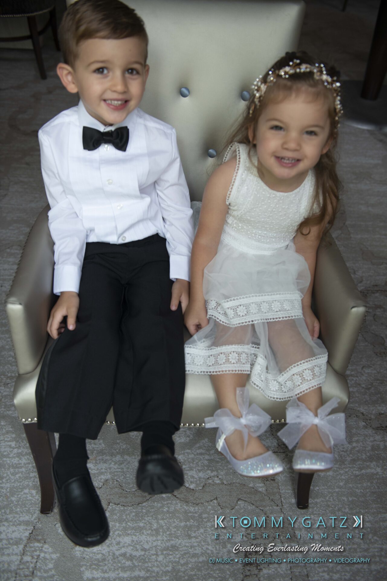 Urania-Aristidis-060218-PREP-Low-Res-WM-57 - TOMMY GATZ ENTERTAINMENT A young boy in a tuxedo and a girl in a white dress sit together on a silver chair, smiling.