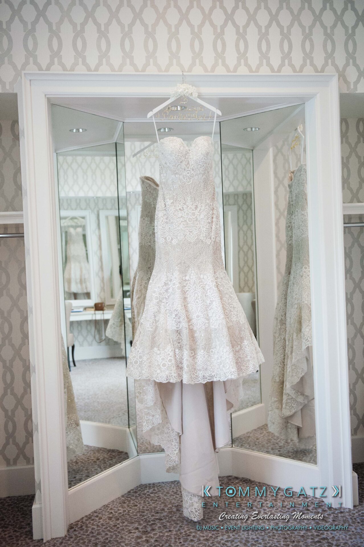 Urania-Aristidis-060218-PREP-Low-Res-WM-6 - TOMMY GATZ ENTERTAINMENT Strapless lace wedding gown hanging in front of a mirrored wall with a patterned wallpaper background.
