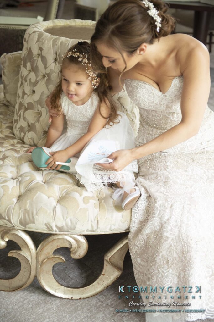 A woman in a wedding dress sits with a young girl in a white dress on a tufted sofa. The girl holds a blue hairbrush.