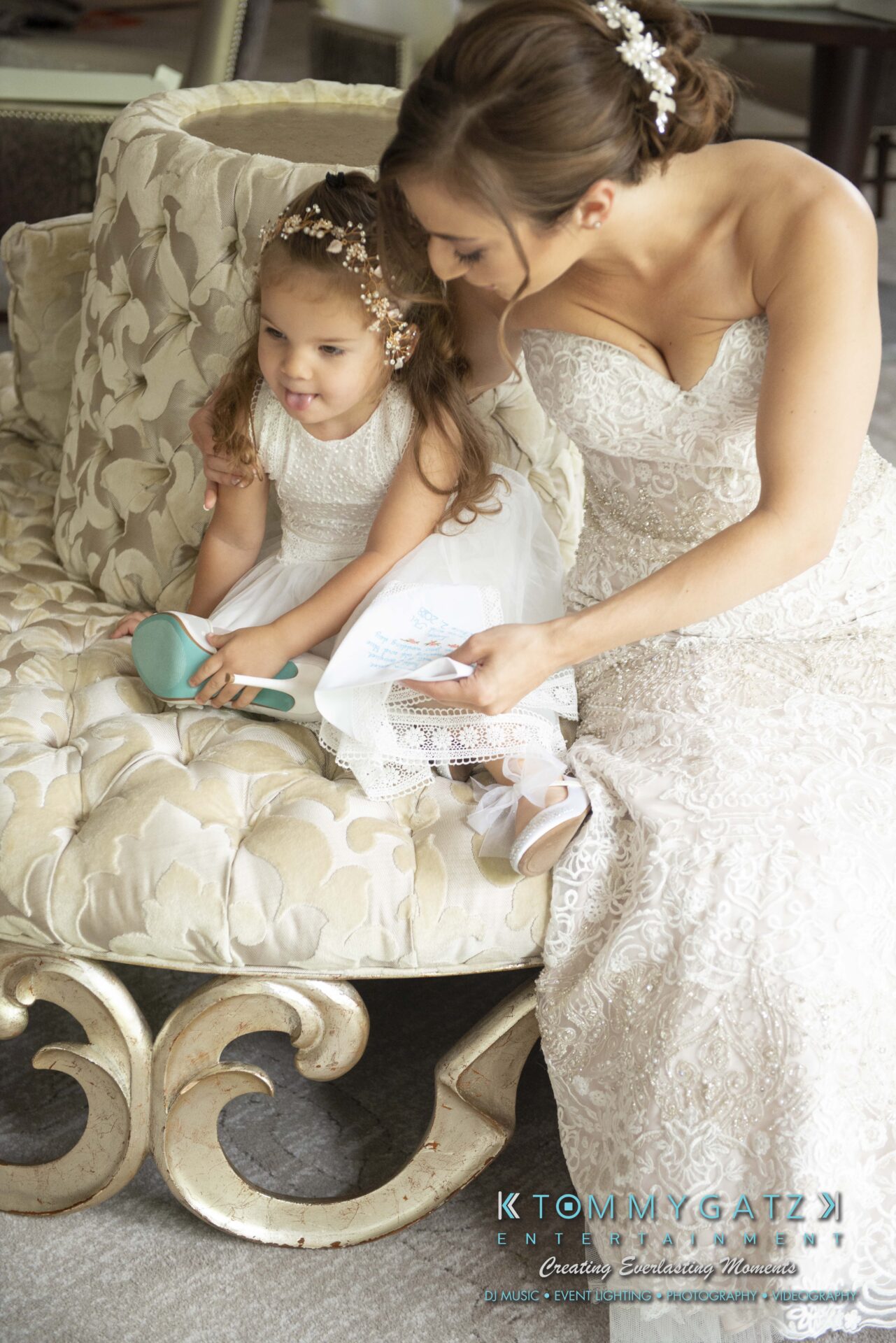 Urania-Aristidis-060218-PREP-Low-Res-WM-61 - TOMMY GATZ ENTERTAINMENT A bride and a young girl, both in white dresses, sit on a plush chair. The girl is holding a blue hairbrush and wearing floral hair accessories. The bride has her hair styled up.