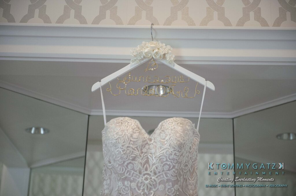 Urania-Aristidis-060218-PREP-Low-Res-WM-7 - TOMMY GATZ ENTERTAINMENT A lace wedding dress on a personalized hanger with the date and names in wire, reflected in a mirror, under a patterned ceiling.