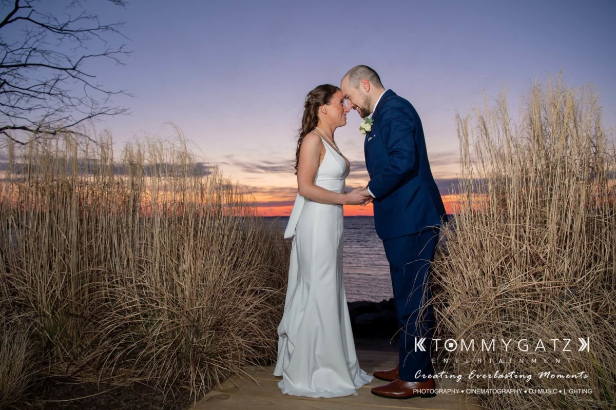 BrittanyDaniel-WEDDING-Highlights-100 - TOMMY GATZ ENTERTAINMENT A couple in wedding attire stands close together, holding hands, with a sunset over a body of water in the background.