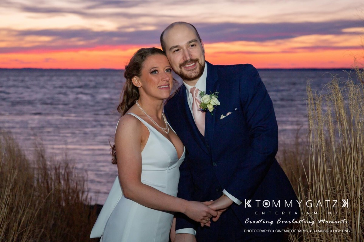 BrittanyDaniel-WEDDING-Highlights-101 - TOMMY GATZ ENTERTAINMENT A couple in formal attire poses by a lake during sunset, surrounded by tall grass.