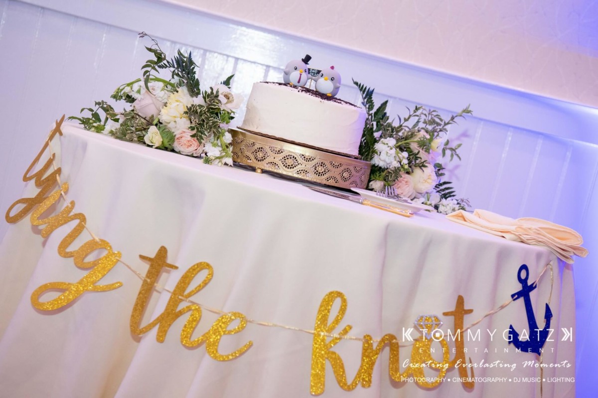 BrittanyDaniel-WEDDING-Highlights-102 - TOMMY GATZ ENTERTAINMENT Wedding cake on a decorated table with floral arrangements and a "tying the knot" sign.