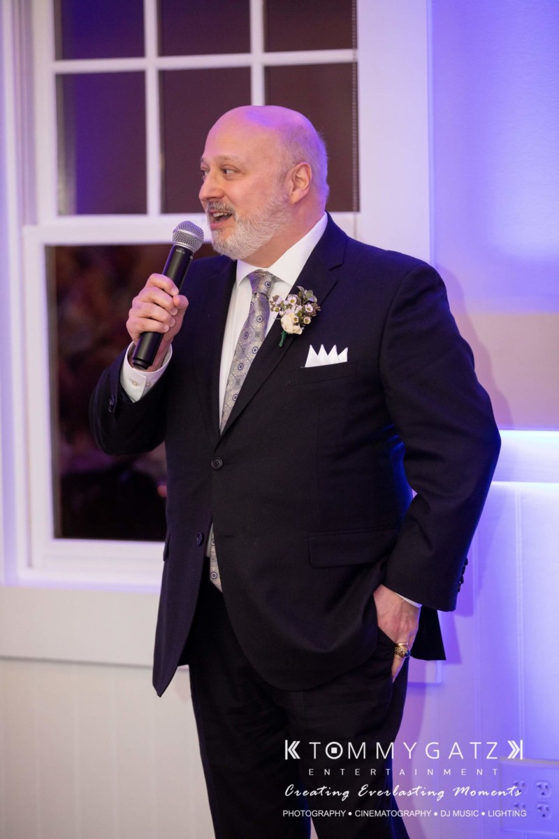 BrittanyDaniel-WEDDING-Highlights-104 - TOMMY GATZ ENTERTAINMENT A man in a suit speaks into a microphone at an indoor event.