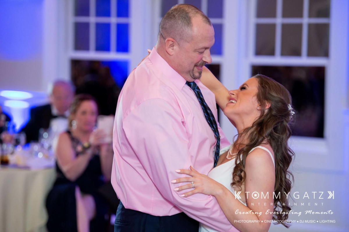 BrittanyDaniel-WEDDING-Highlights-105 - TOMMY GATZ ENTERTAINMENT A couple dances and smiles at an indoor event, with guests seated in the background.