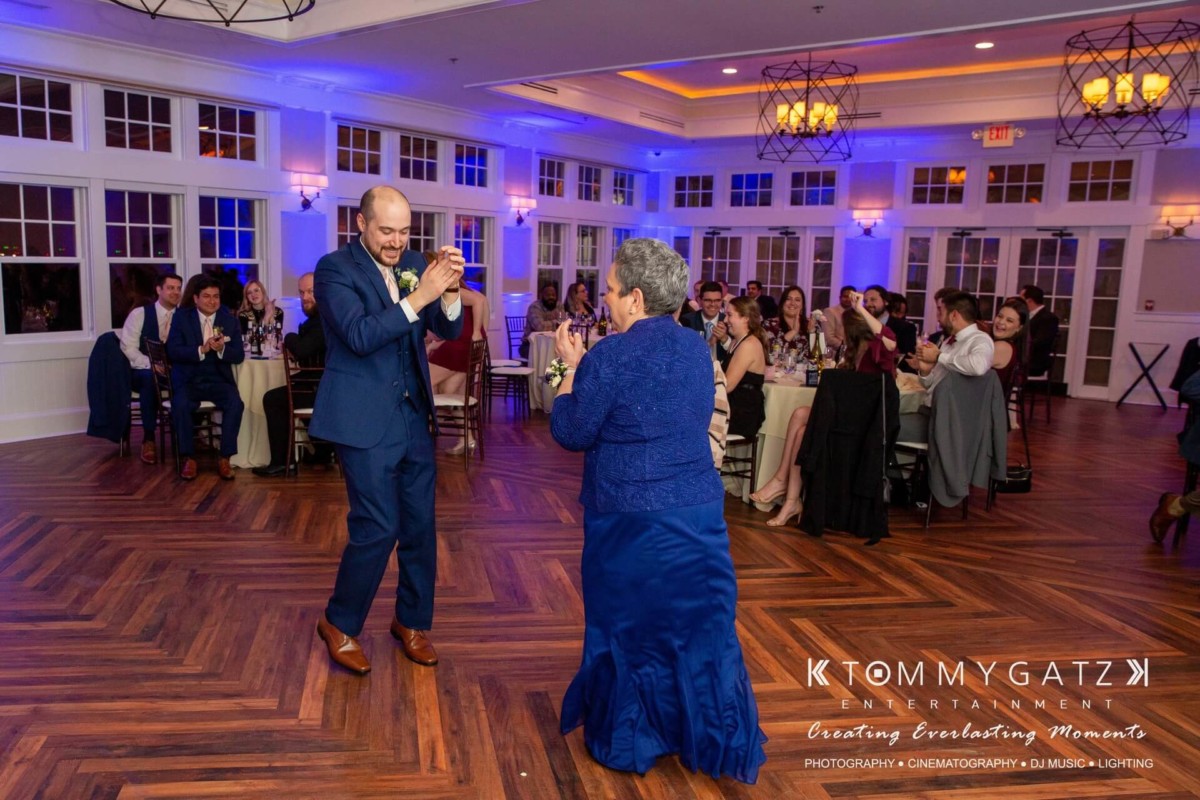 BrittanyDaniel-WEDDING-Highlights-106 - TOMMY GATZ ENTERTAINMENT A man and woman in formal attire are dancing joyfully on a wooden floor in a banquet hall, with guests seated at tables in the background.