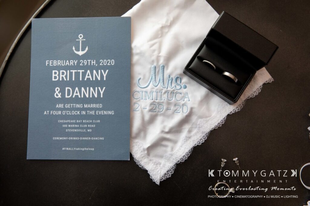 BrittanyDaniel-WEDDING-Highlights-13 - TOMMY GATZ ENTERTAINMENT Wedding invitation and accessories including a handkerchief with "Mrs. Cimiluca 2-29-20" embroidered, two rings in a box, and promotional materials for Tommy Gatz Entertainment.
