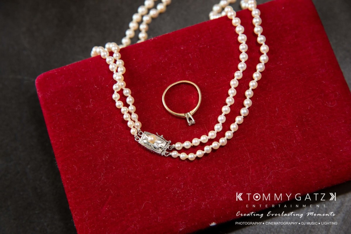 BrittanyDaniel-WEDDING-Highlights-14 - TOMMY GATZ ENTERTAINMENT A pearl necklace and a gold ring on a red velvet surface.