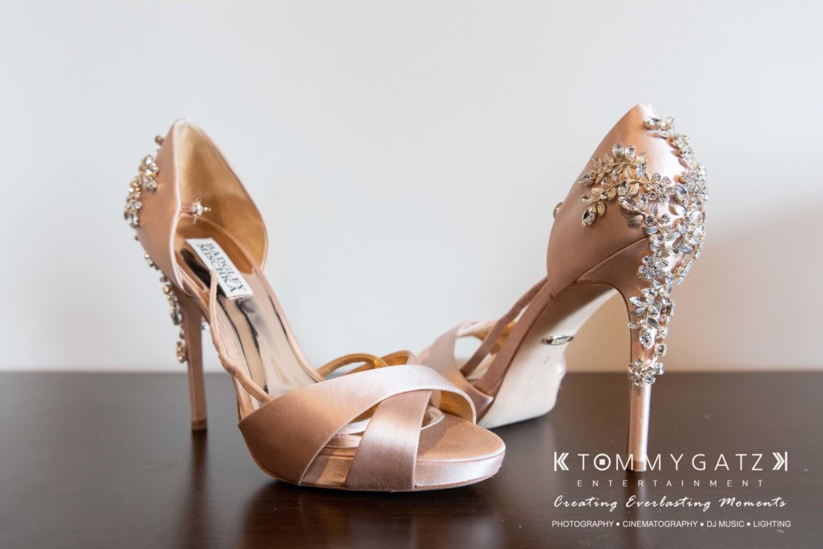 BrittanyDaniel-WEDDING-Highlights-15 - TOMMY GATZ ENTERTAINMENT Elegant high-heeled shoes adorned with rhinestones on a wooden surface. Tommy Gatz Entertainment logo in the corner.