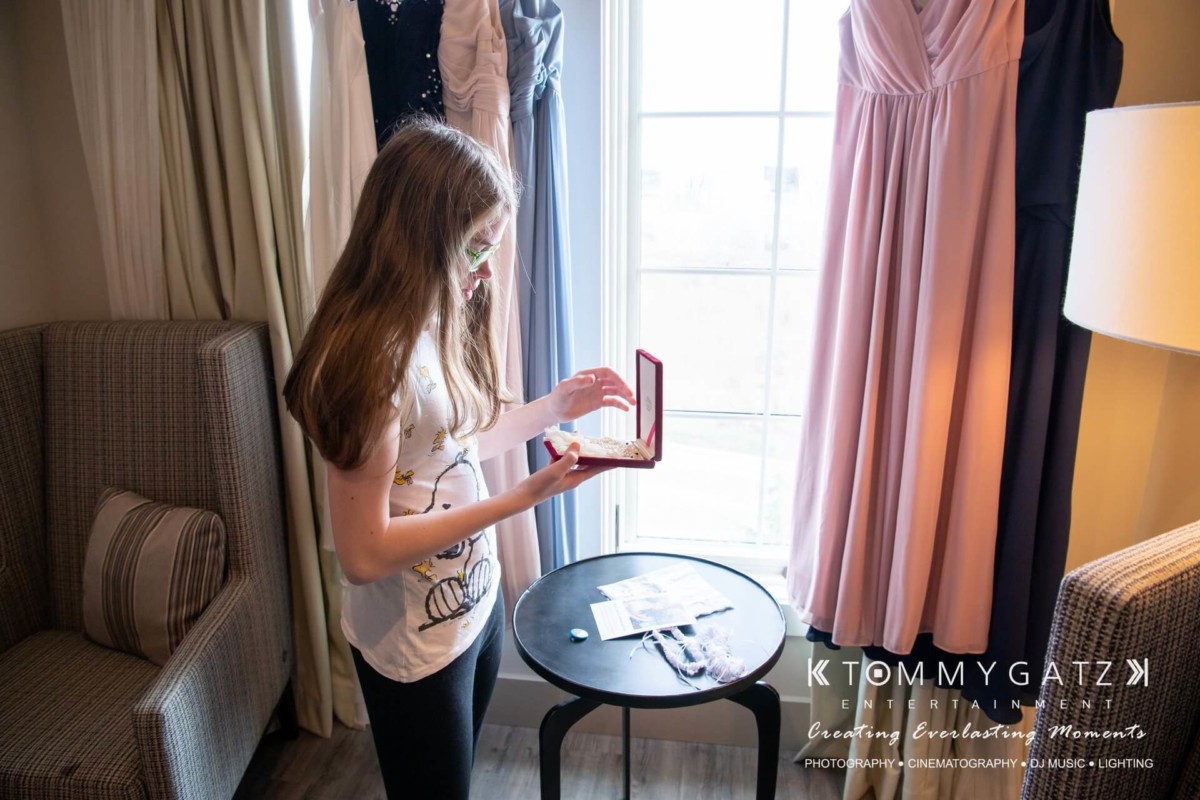 BrittanyDaniel-WEDDING-Highlights-18 - TOMMY GATZ ENTERTAINMENT A young woman stands by a window, holding an open jewelry box. Dresses hang nearby. The scene appears to be part of a preparation or dressing area.