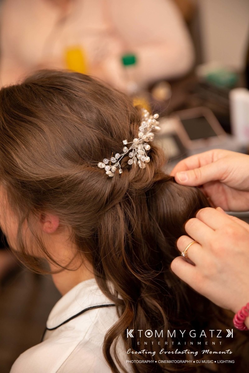 BrittanyDaniel-WEDDING-Highlights-2 - TOMMY GATZ ENTERTAINMENT Person styling a woman's hair, adorned with a decorative hairpiece, during a formal preparation.