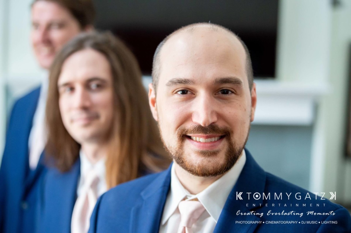 BrittanyDaniel-WEDDING-Highlights-22 - TOMMY GATZ ENTERTAINMENT Three men in suits smiling, with one in focus while the others are blurred in the background.
