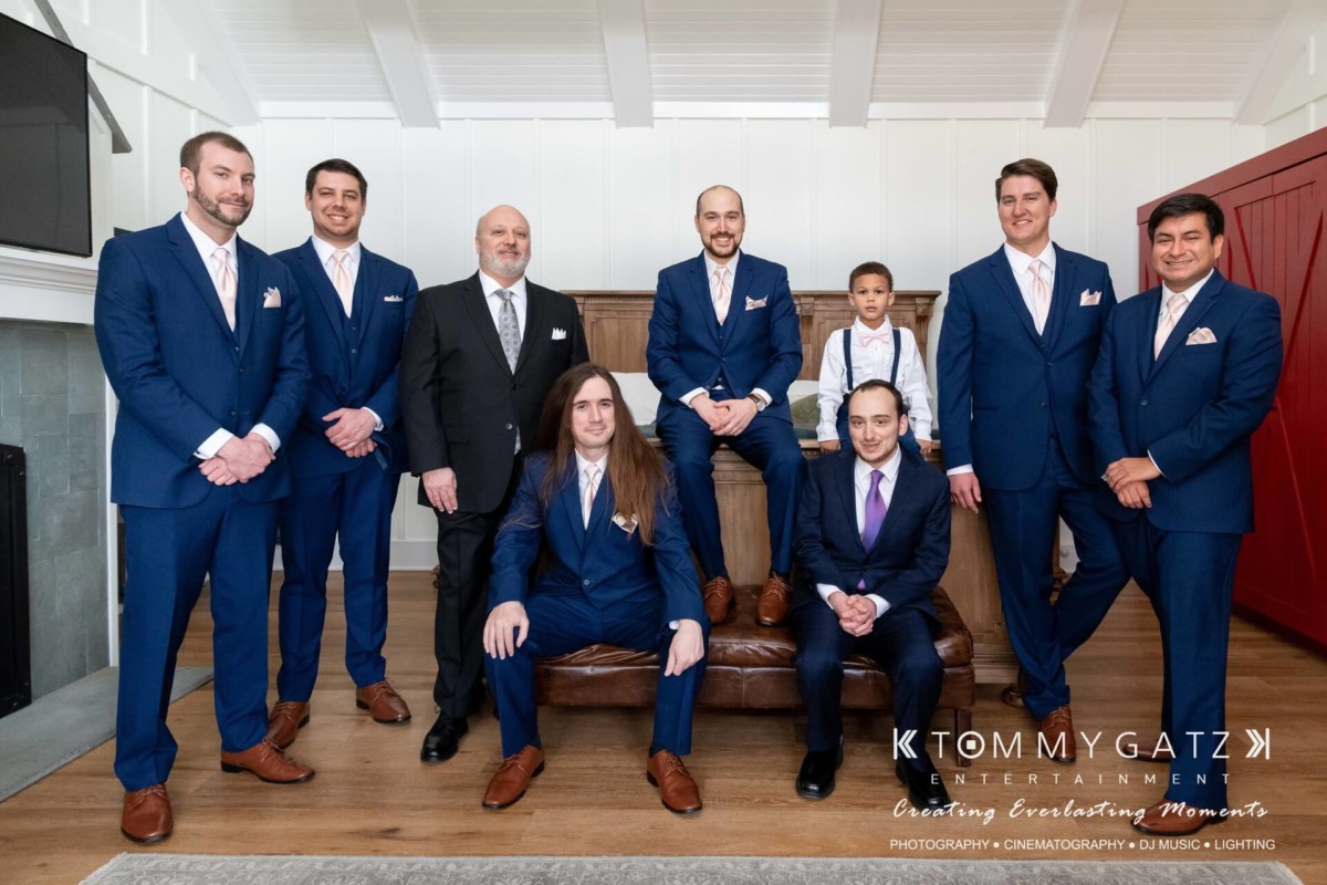 BrittanyDaniel-WEDDING-Highlights-23 - TOMMY GATZ ENTERTAINMENT A group of nine men in formal attire pose indoors, some seated and others standing. The setting includes a fireplace and red barn-style doors.