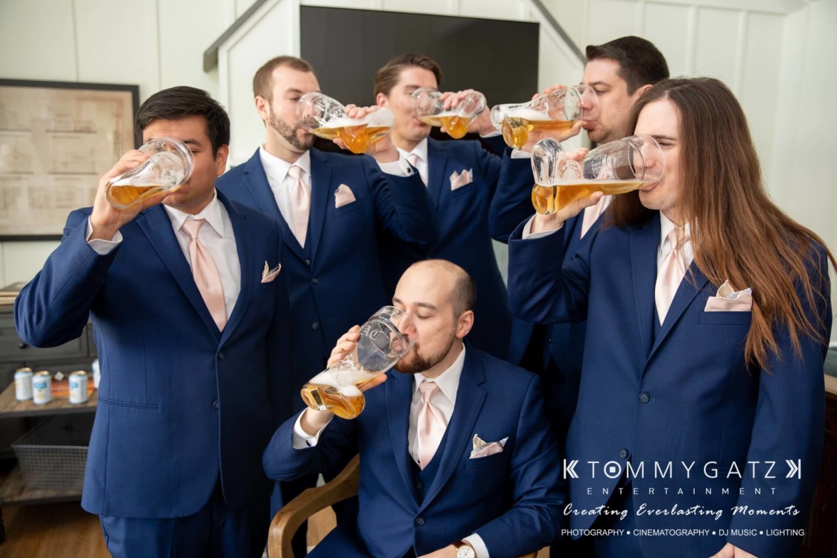 BrittanyDaniel-WEDDING-Highlights-24 - TOMMY GATZ ENTERTAINMENT A group of six men in blue suits drink beer from large mugs indoors.
