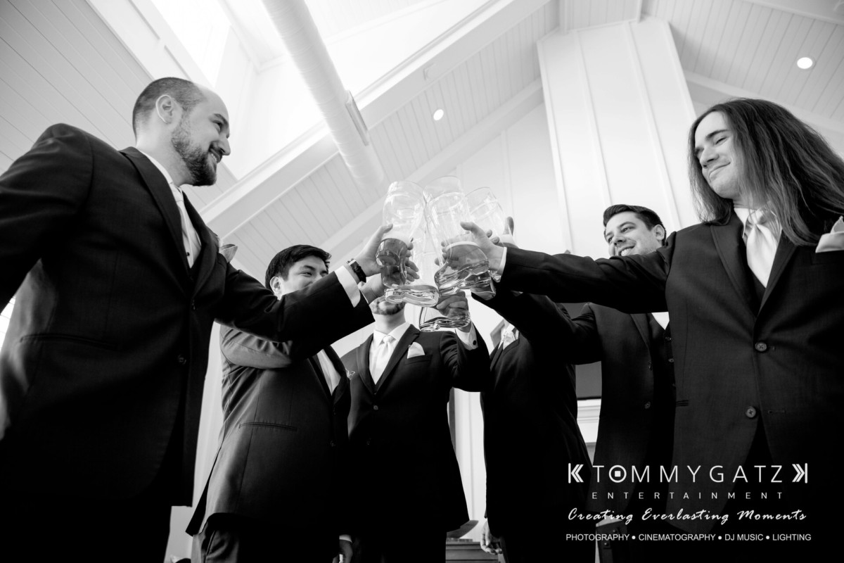BrittanyDaniel-WEDDING-Highlights-25 - TOMMY GATZ ENTERTAINMENT A group of men in suits clink glasses in a celebratory toast under a high ceiling.
