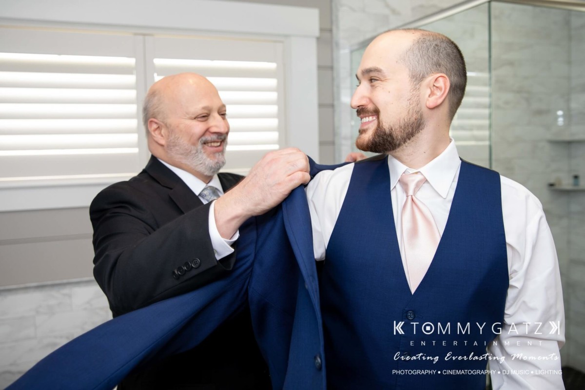 BrittanyDaniel-WEDDING-Highlights-28 - TOMMY GATZ ENTERTAINMENT A man helps another man put on a suit jacket in a well-lit room with white shutters and a marble wall.