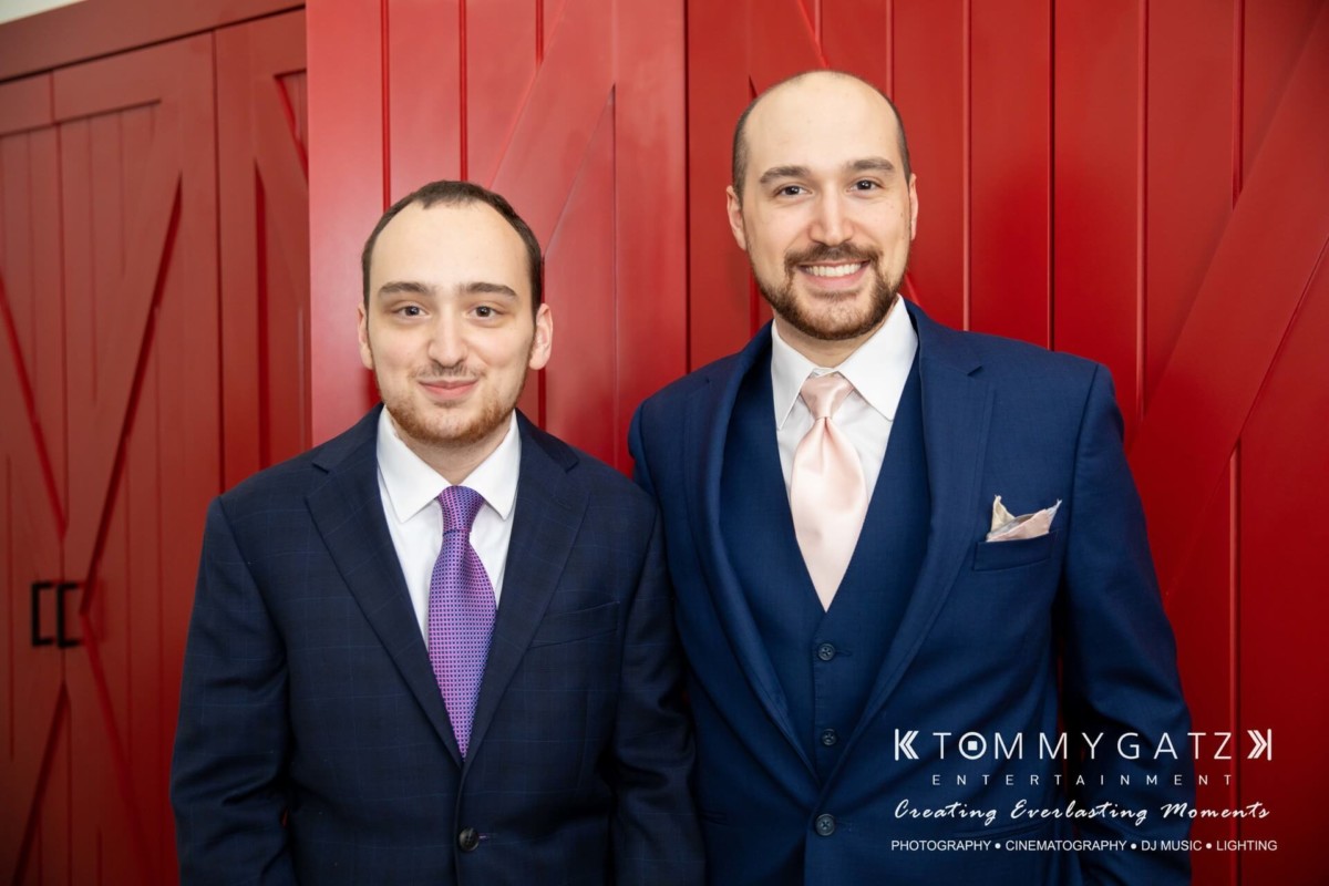 BrittanyDaniel-WEDDING-Highlights-29 - TOMMY GATZ ENTERTAINMENT Two men in suits stand in front of a red wall. One is wearing a dark blue suit with a purple tie, and the other a lighter blue suit with a pink tie.
