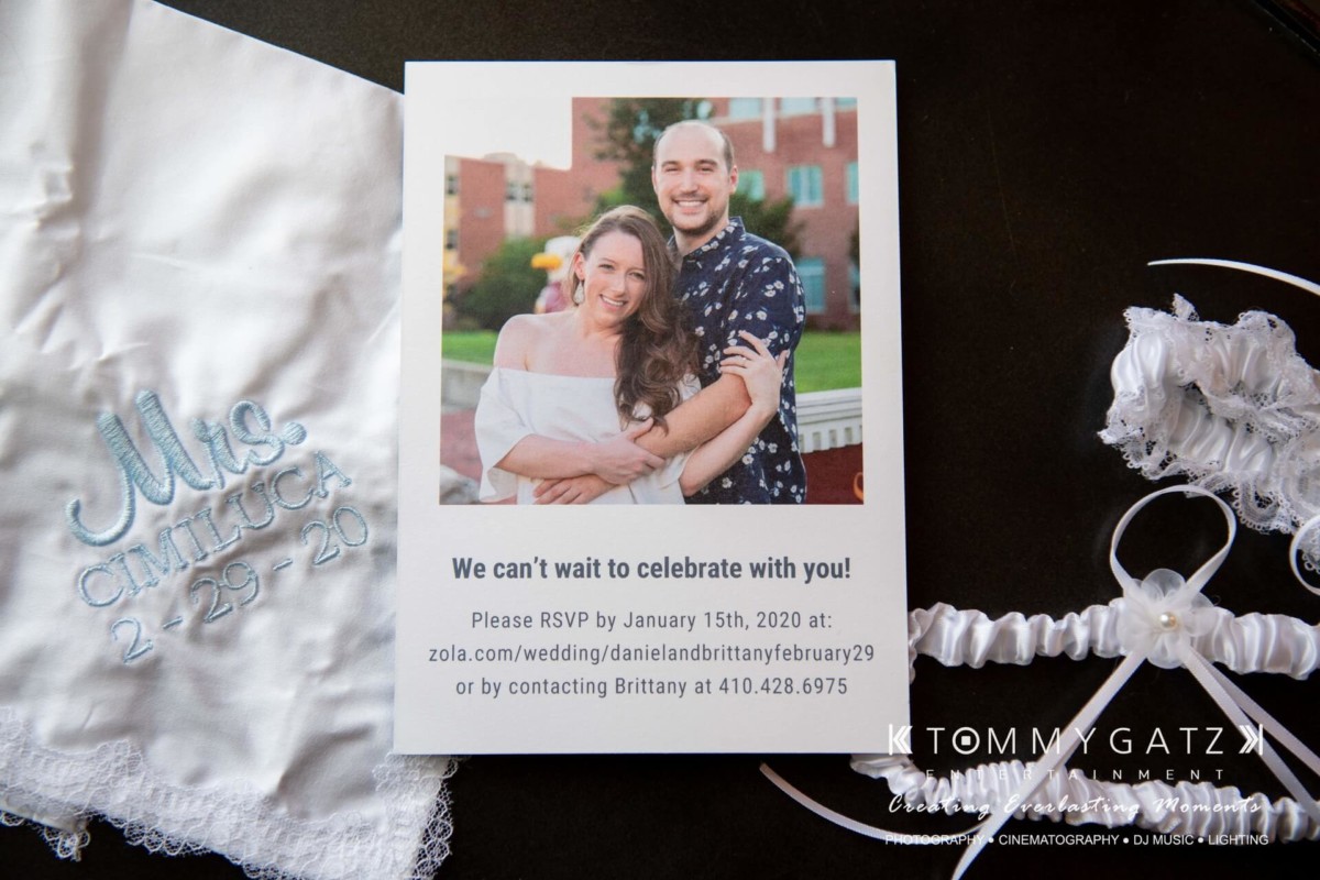 BrittanyDaniel-WEDDING-Highlights-30 - TOMMY GATZ ENTERTAINMENT Wedding invitation with a smiling couple's photo. Text includes RSVP instructions and date "2-29-20." Embroidered handkerchief reads "Mr. & Mrs. Cimilluca 2-29-20" beside a garter.