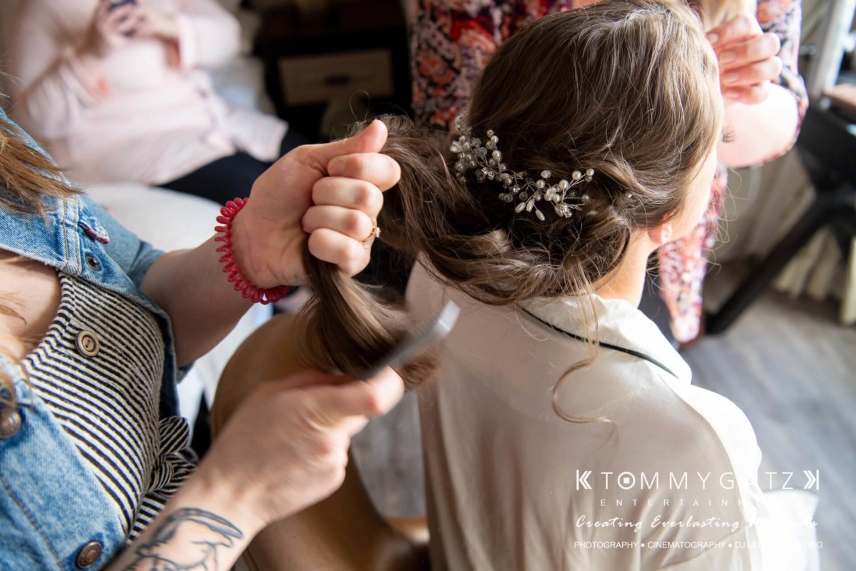 BrittanyDaniel-WEDDING-Highlights-35 - TOMMY GATZ ENTERTAINMENT A person styles another's hair, using a comb and hairpin with floral details, while seated indoors.