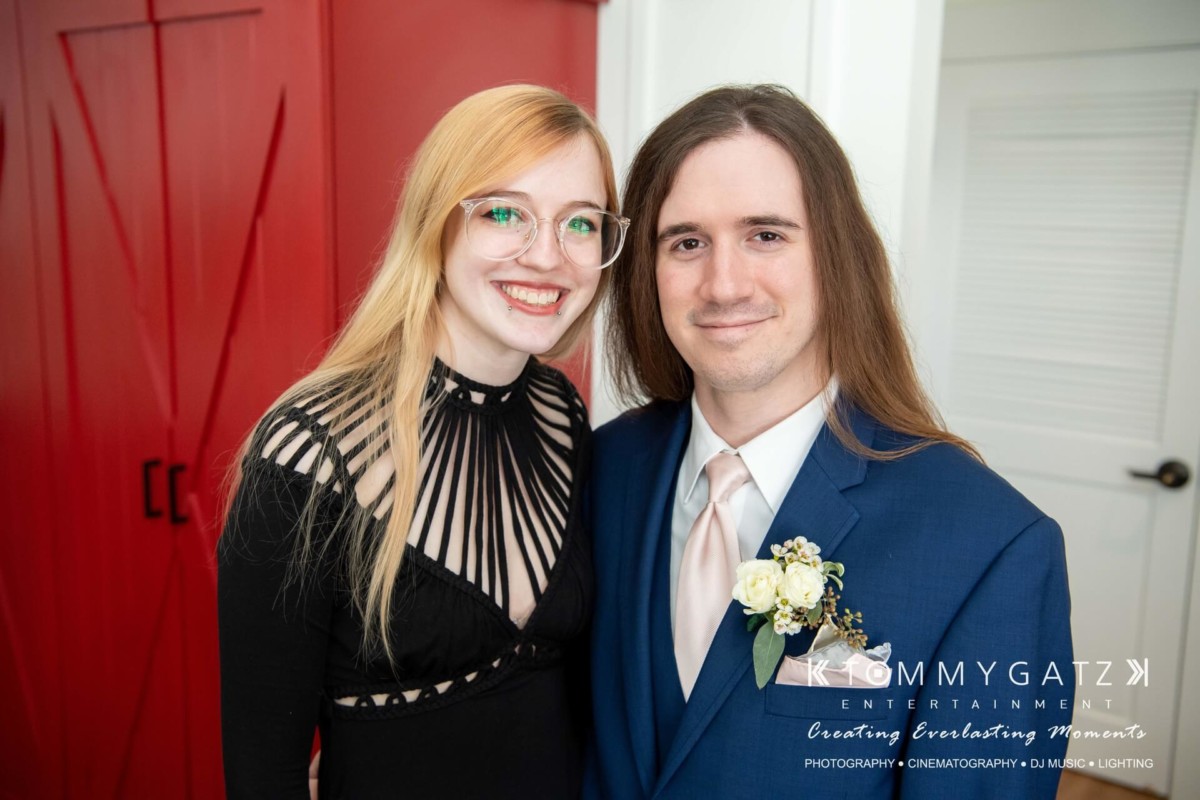 BrittanyDaniel-WEDDING-Highlights-41 - TOMMY GATZ ENTERTAINMENT A woman in a black dress and a man in a blue suit with a boutonniere stand smiling in front of red double doors.