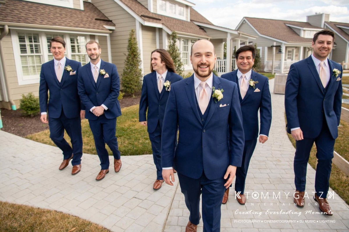 BrittanyDaniel-WEDDING-Highlights-42 - TOMMY GATZ ENTERTAINMENT A groom and five groomsmen in matching blue suits and pink ties walk outside on a stone path near houses.