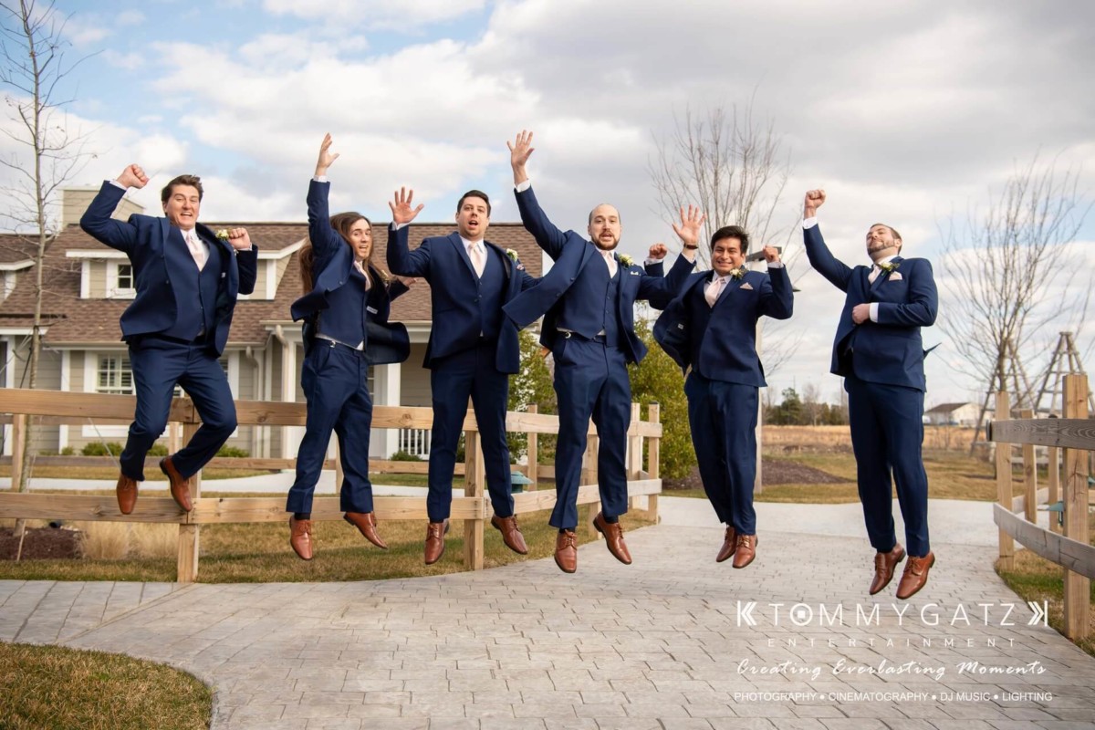 BrittanyDaniel-WEDDING-Highlights-44 - TOMMY GATZ ENTERTAINMENT Six men in blue suits jump joyfully on a paved pathway in front of a house, with a clear sky and trees in the background.