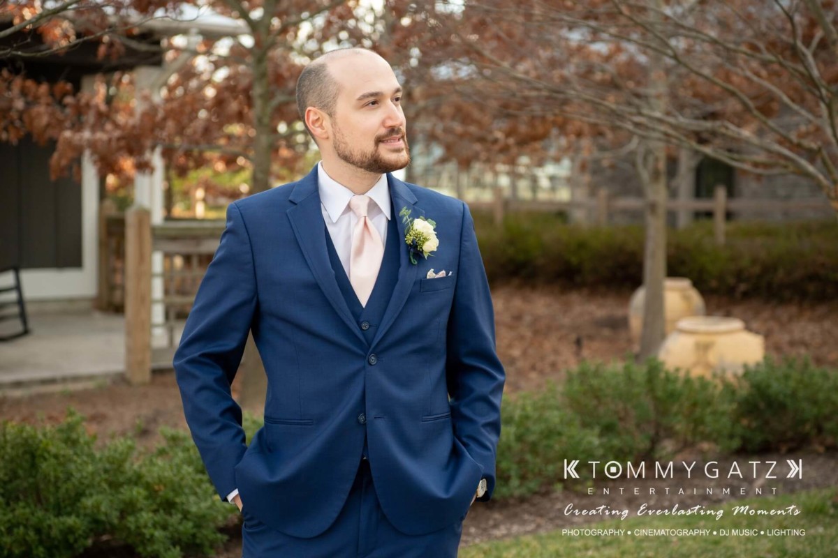 BrittanyDaniel-WEDDING-Highlights-45 - TOMMY GATZ ENTERTAINMENT A man in a blue suit with a boutonniere stands outdoors, looking to the side. Behind him are trees and a wooden fence. The image has a logo for Tommy Gatz Entertainment.