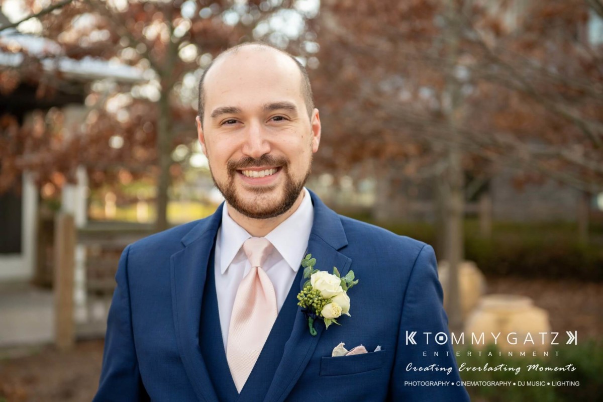 BrittanyDaniel-WEDDING-Highlights-46 - TOMMY GATZ ENTERTAINMENT A man in a blue suit with a pink tie and boutonniere smiles outdoors. The background shows blurred trees and a building.