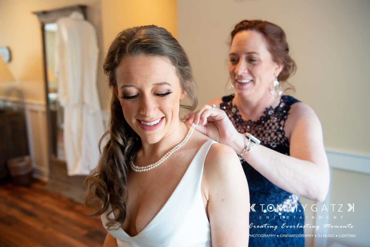BrittanyDaniel-WEDDING-Highlights-47 - TOMMY GATZ ENTERTAINMENT A woman helps another woman put on a necklace in a well-lit room. They both appear happy.