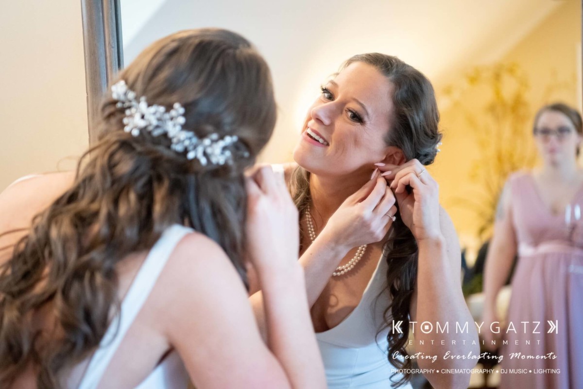 BrittanyDaniel-WEDDING-Highlights-48 - TOMMY GATZ ENTERTAINMENT A woman in a white dress is adjusting an earring while looking into a mirror. Another woman in a pink dress is visible in the background.