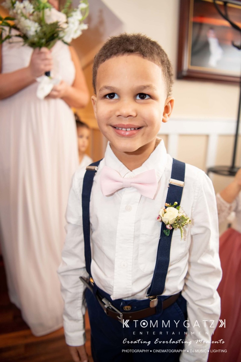 BrittanyDaniel-WEDDING-Highlights-50 - TOMMY GATZ ENTERTAINMENT A smiling boy in a white shirt, pink bow tie, and navy suspenders stands indoors. He is wearing a boutonniere. A person in the background holds flowers.