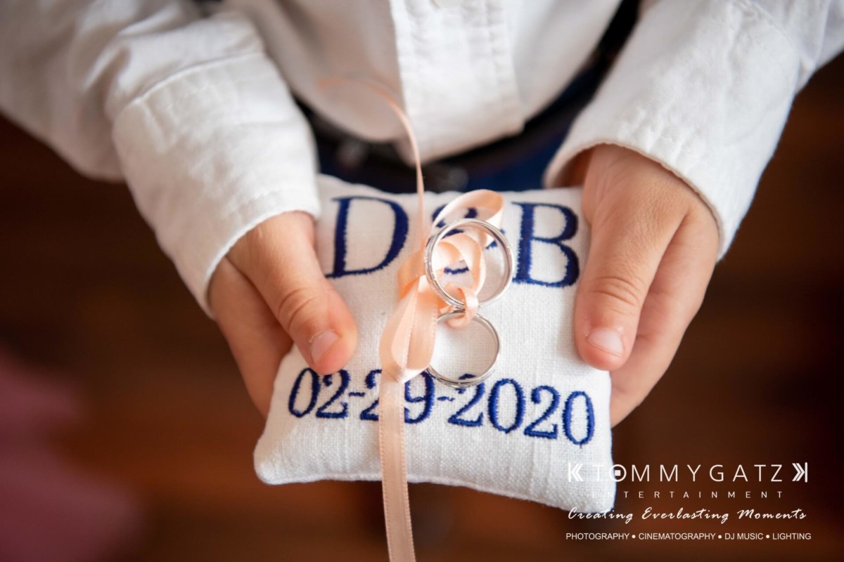 BrittanyDaniel-WEDDING-Highlights-51 - TOMMY GATZ ENTERTAINMENT Child holding a white pillow with rings tied by a ribbon, embroidered with initials "D & B" and date "02-29-2020".