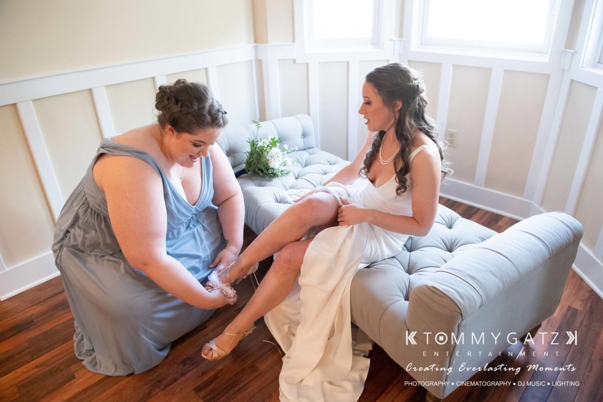 BrittanyDaniel-WEDDING-Highlights-54 - TOMMY GATZ ENTERTAINMENT A bridesmaid helps a bride adjust her shoe while sitting on a gray couch in a well-lit room.
