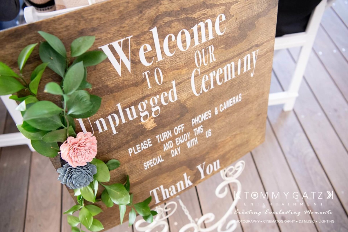 BrittanyDaniel-WEDDING-Highlights-56 - TOMMY GATZ ENTERTAINMENT A wooden sign with the text "Welcome to our unplugged ceremony" asking guests to turn off phones and cameras. Decorated with flowers and leaves.