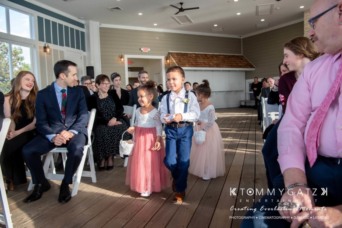 BrittanyDaniel-WEDDING-Highlights-57 - TOMMY GATZ ENTERTAINMENT Children dressed formally walk down the aisle carrying flowers at an indoor wedding ceremony, with guests seated on either side.