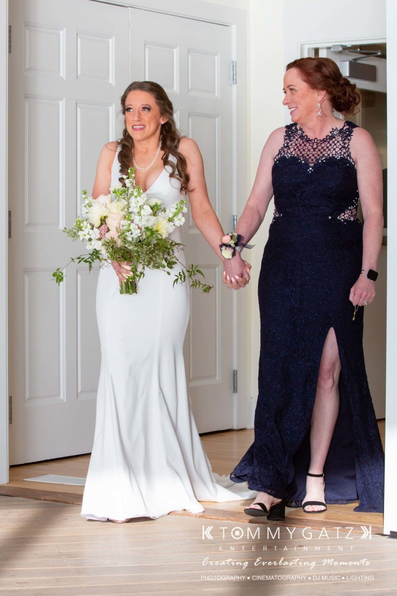BrittanyDaniel-WEDDING-Highlights-58 - TOMMY GATZ ENTERTAINMENT A bride in a white dress and a woman in a navy gown holding a bouquet walk arm-in-arm through a doorway.