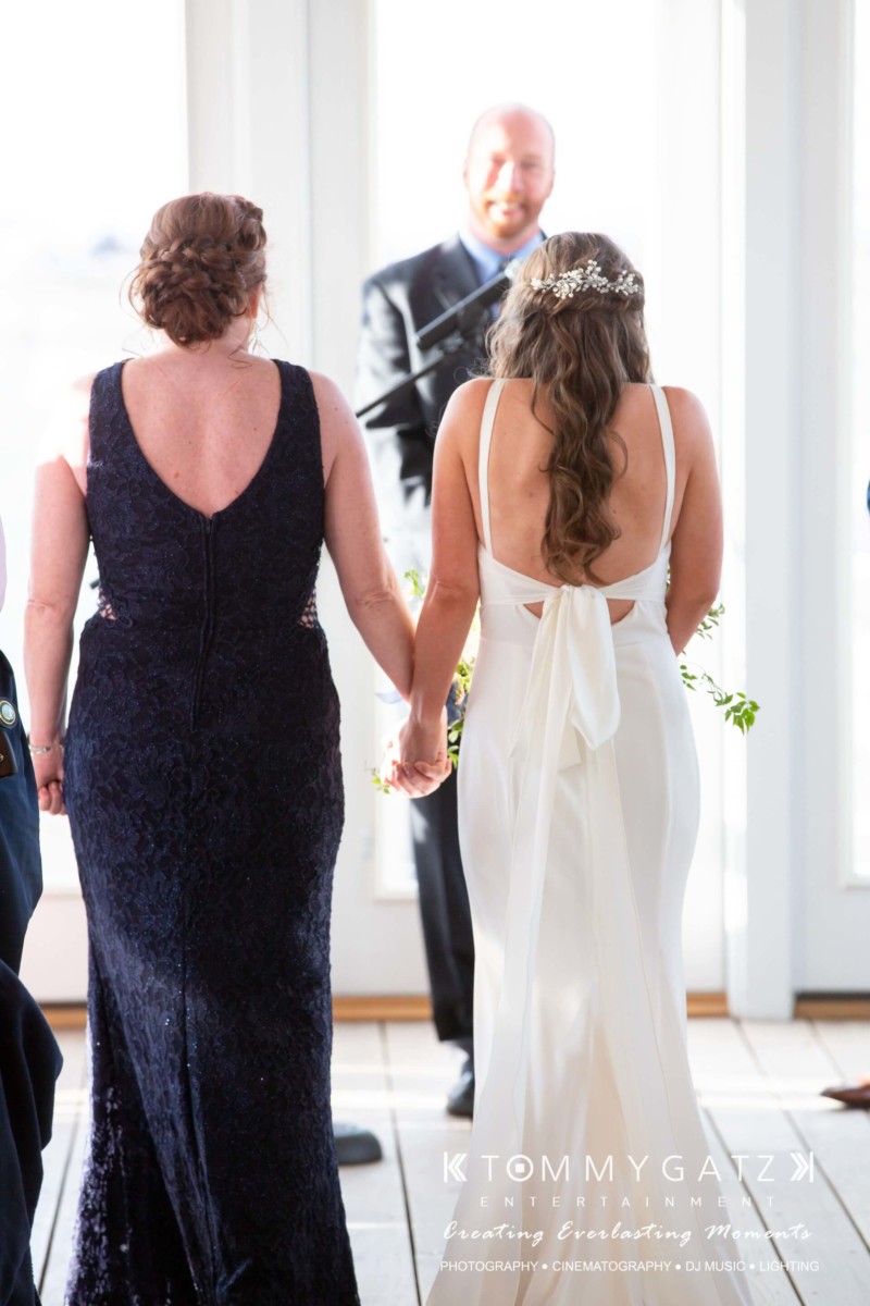 BrittanyDaniel-WEDDING-Highlights-59 - TOMMY GATZ ENTERTAINMENT Two women, one in a dark blue dress and the other in a white gown, hold hands and walk towards a man standing in front of a door.