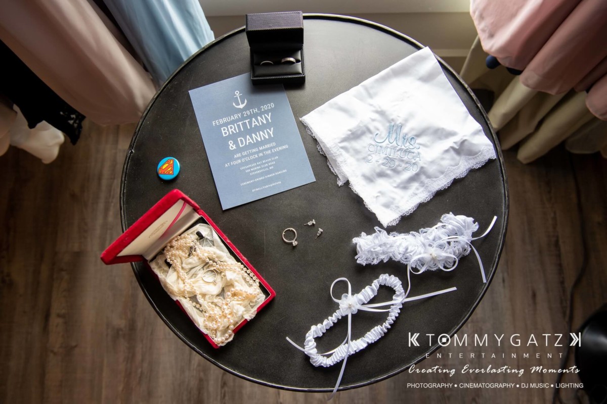 BrittanyDaniel-WEDDING-Highlights-6 - TOMMY GATZ ENTERTAINMENT Wedding preparation items on a round table: invitation card, embroidered handkerchief, garter, jewelry, and a ring box.