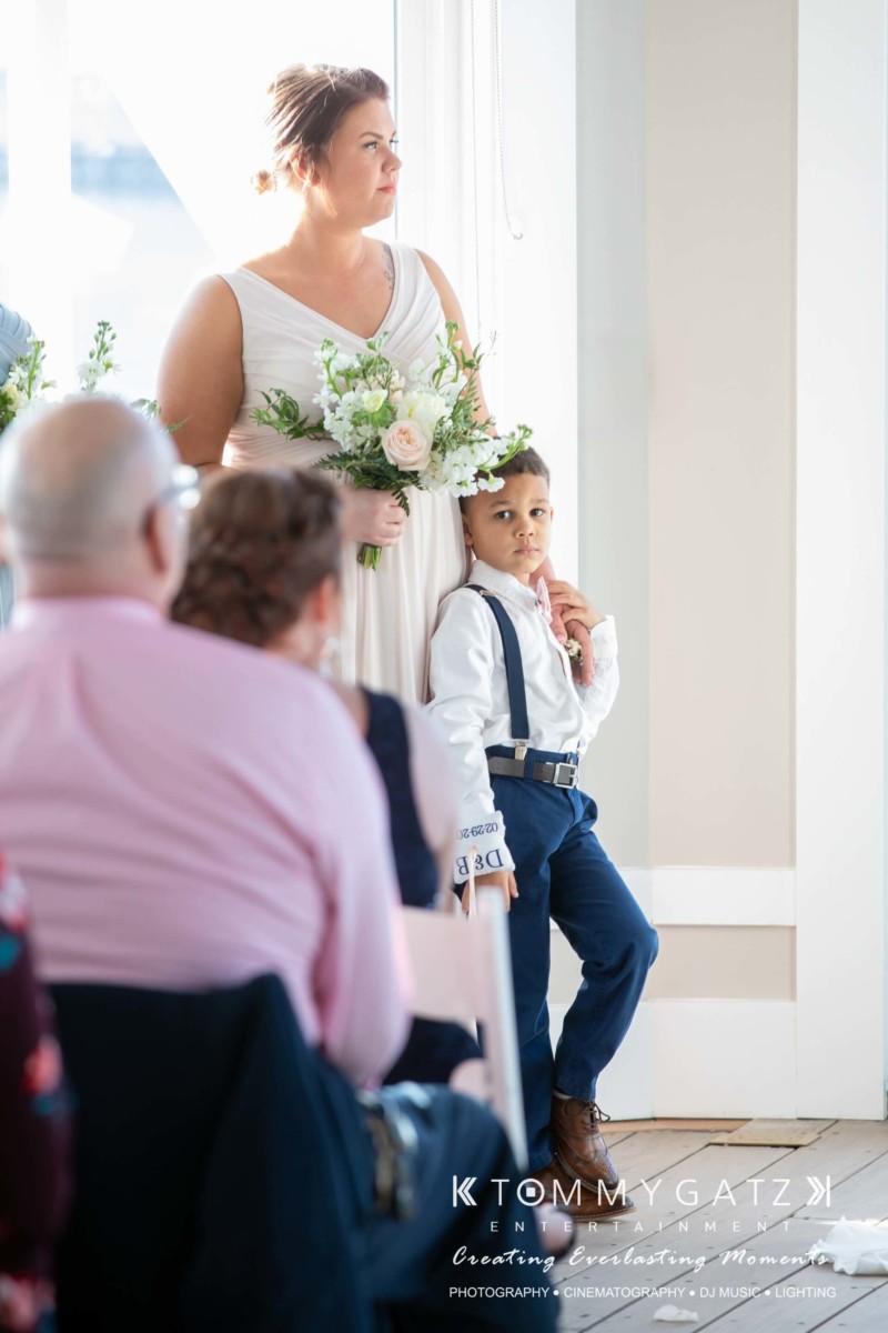 BrittanyDaniel-WEDDING-Highlights-60 - TOMMY GATZ ENTERTAINMENT A woman in a white dress holds a bouquet while a boy in a white shirt and blue suspenders stands beside her, next to seated guests.
